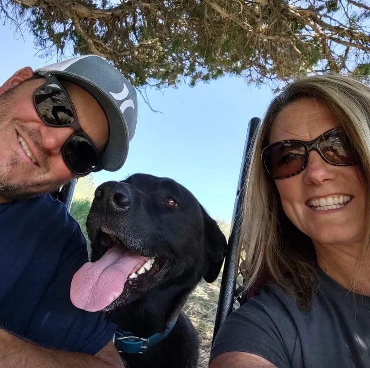 Matt and Nichole smiling in sunglasses outdoors with their black Lab between them